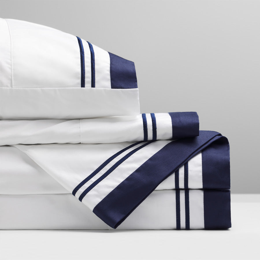 Stacked Manhattan Luxury Bedding sheets in 400TC GOTS-certified cotton sateen, featuring a crisp white base, navy header, and satin-stitch embroidery.